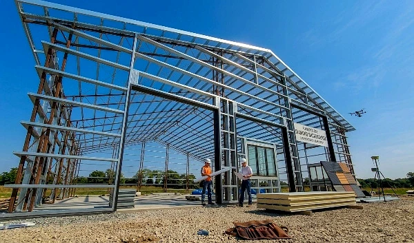 A wide-angle view of a large modern barndominium under construction featuring a heavy-duty steel frame, open bays for oversized doors, and architectural plans laid out in the foreground on a gravel construction site.