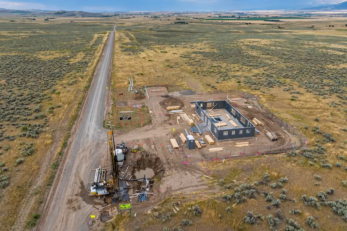 Aerial view of a barndominium under construction on rural Idaho land, showing steel framing, a well drilling rig, and septic site preparation